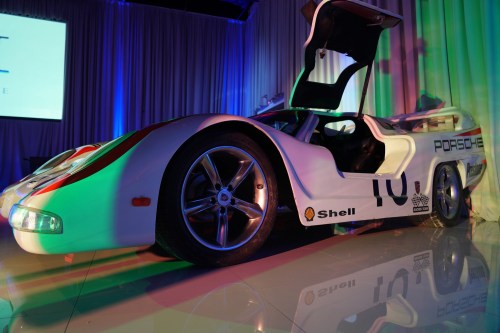 A sleek, white Porsche race car with an open door, illuminated by colorful lights in a stylish setting.
