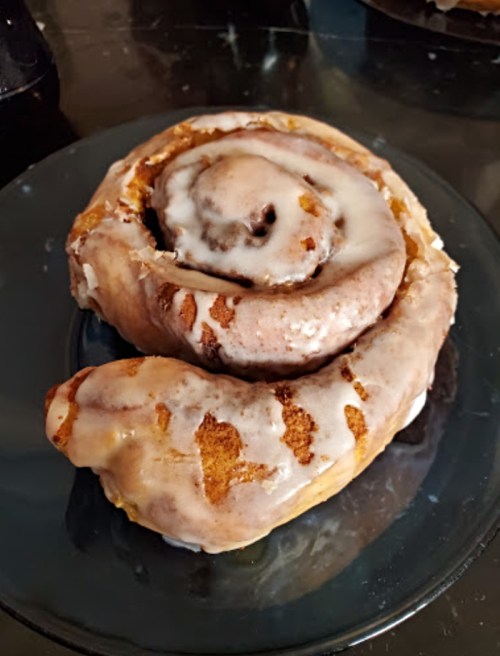 A large, gooey cinnamon roll with icing, sitting on a black plate against a dark background.
