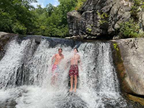 Two boys stand in a waterfall, splashing water around them, surrounded by lush greenery and rocky terrain.