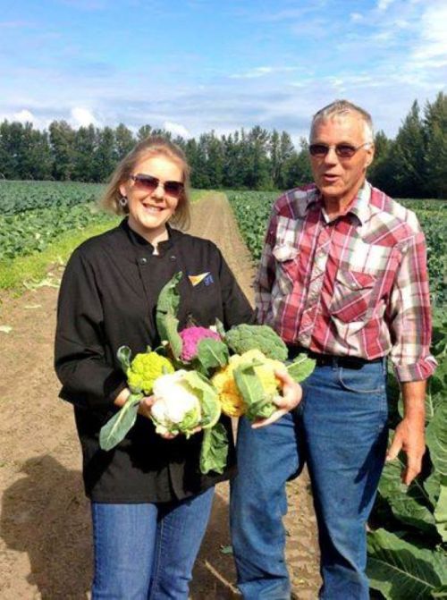 A woman in a black jacket and a man in a plaid shirt stand in a field, holding fresh vegetables.
