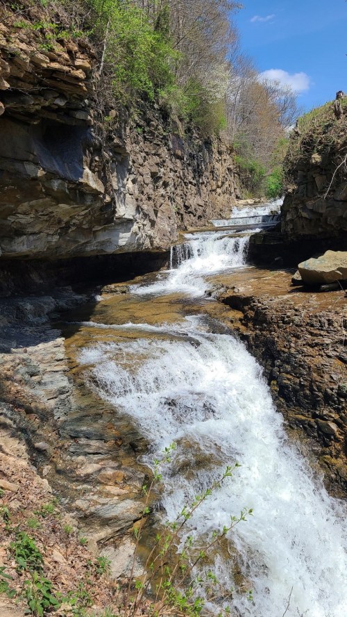 A serene waterfall cascades over rocky terrain, surrounded by lush greenery and a clear blue sky.
