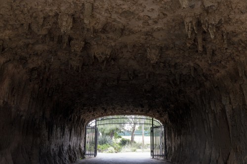 A dark tunnel with textured walls, leading to an open gate and a view of greenery outside.