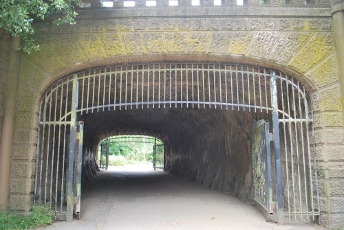 A stone archway with metal gates leads into a dark tunnel surrounded by greenery.