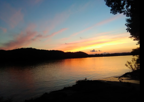 A serene sunset over a calm lake, with colorful clouds reflecting on the water and silhouettes of distant hills.