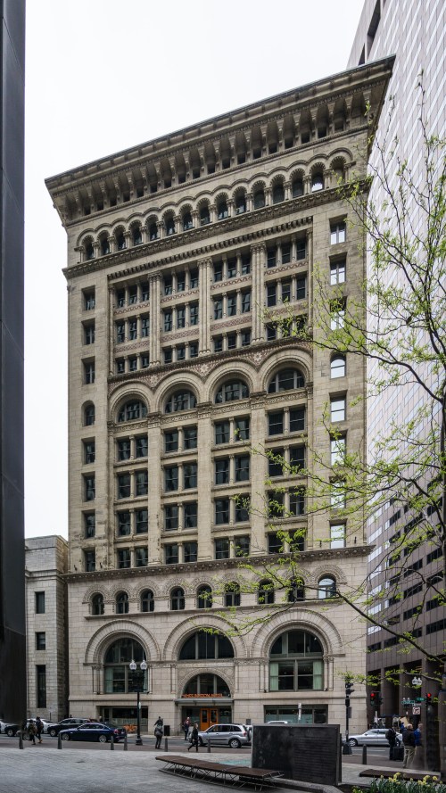 Historic building with ornate architecture, featuring arched windows and a decorative cornice, surrounded by modern skyscrapers.