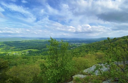 A scenic view of rolling green hills under a blue sky with fluffy clouds, framed by trees and rocky outcrops.