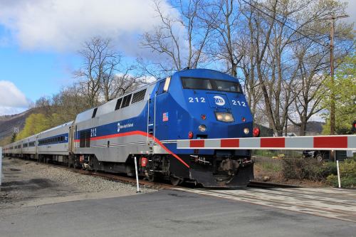 A blue and silver train labeled "212" approaches a level crossing with a barrier down, surrounded by trees and a cloudy sky.