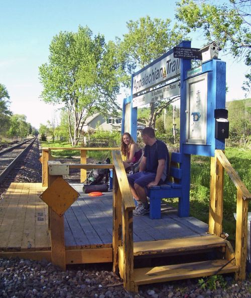 Two people sit on a wooden platform at a train station near the Appalachian Trail, surrounded by greenery.