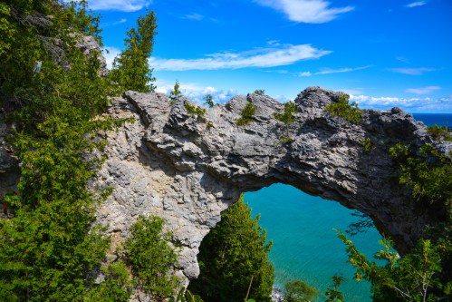 A natural stone arch surrounded by greenery, overlooking a turquoise body of water under a bright blue sky.