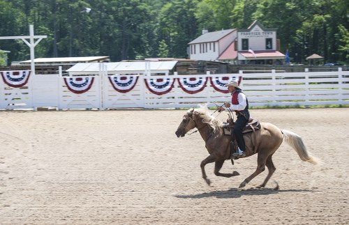 A rider in western attire gallops on a horse in an outdoor arena decorated with red, white, and blue bunting.