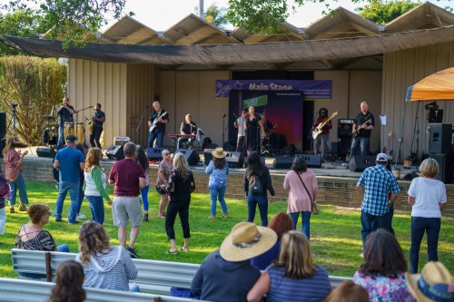 A band performs on stage while an audience dances and enjoys the outdoor concert in a park setting.