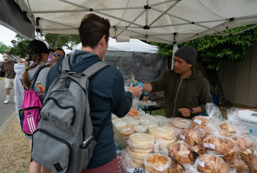 A vendor hands a drink to a customer at a market stall, surrounded by baked goods and other shoppers.
