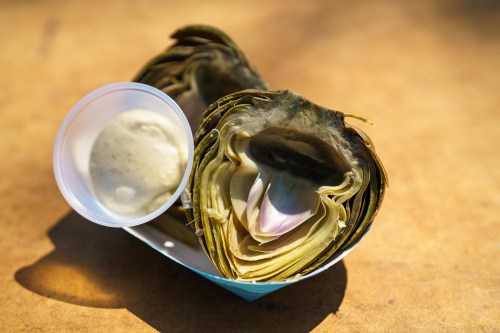 Two halved artichokes served with a small cup of dipping sauce on a brown surface.