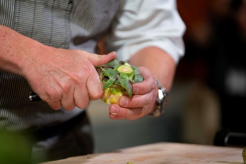 A chef's hands carefully prepare an artichoke, removing the outer leaves with a knife.