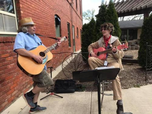 Two musicians play guitars outdoors, smiling and enjoying a performance near a brick building and greenery.