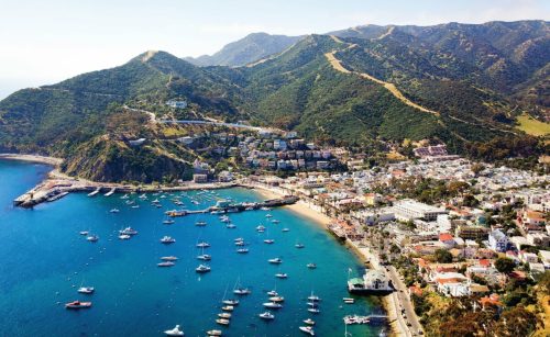 Aerial view of a coastal town with boats in the harbor, surrounded by green hills and mountains.