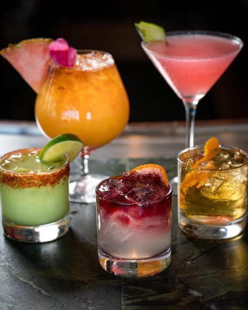 A variety of colorful cocktails on a marble surface, featuring garnishes like fruit and flowers.