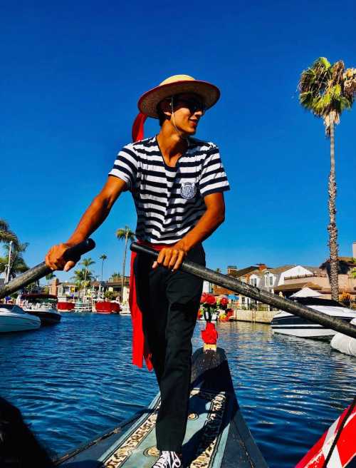 A person in a striped shirt and straw hat rows a boat through a sunny canal lined with palm trees and houses.