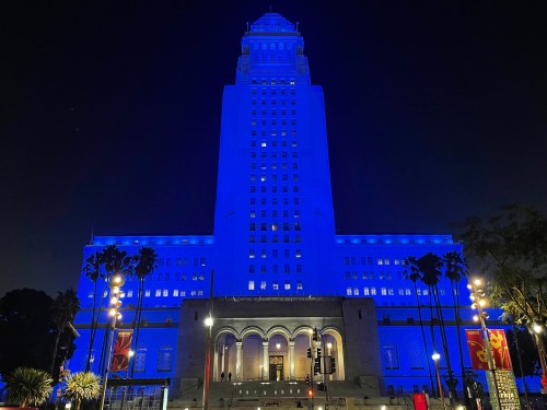 Los Angeles City Hall illuminated in blue at night, surrounded by palm trees and streetlights.