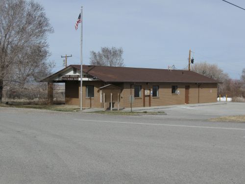 A small brown building with a flagpole, located at a road intersection, surrounded by sparse trees and open land.