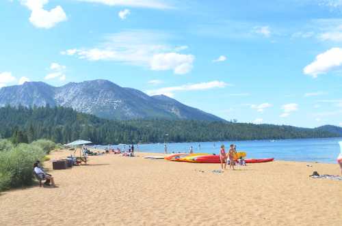 A sandy beach by a lake with people sunbathing, kayaking, and mountains in the background under a blue sky.