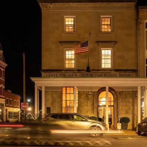 Historic bank building at night, featuring large windows and an American flag, with car lights streaking by.