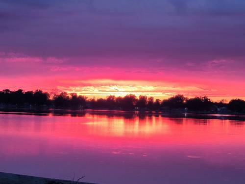 Vibrant sunset over a calm lake, with pink and orange hues reflecting on the water and silhouetted trees in the background.