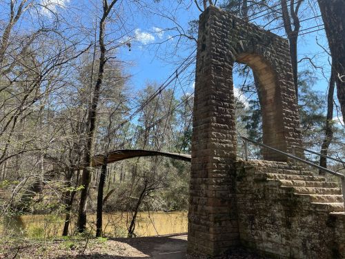 Stone archway with a wooden bridge over a river, surrounded by trees under a clear blue sky.