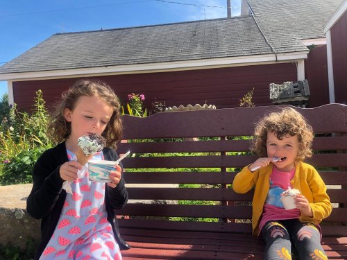 Two children sitting on a bench enjoying ice cream on a sunny day, with a red barn in the background.