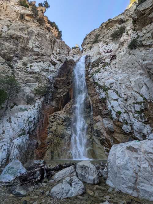 A tall waterfall cascading down rocky cliffs, surrounded by greenery and boulders under a clear blue sky.