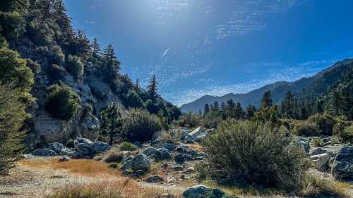 A scenic landscape featuring rocky terrain, lush greenery, and mountains under a bright blue sky with scattered clouds.