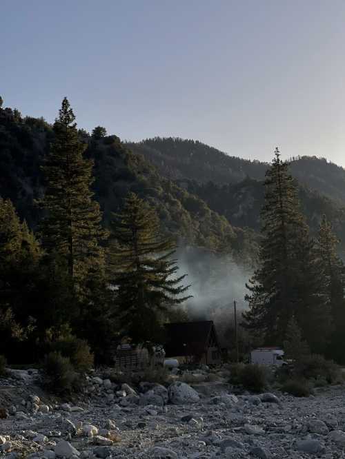 A serene mountain landscape with tall trees, a cabin, and a hint of smoke rising in the distance.