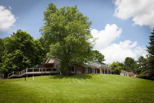 A modern house on a grassy hill, surrounded by trees and under a blue sky with fluffy clouds.
