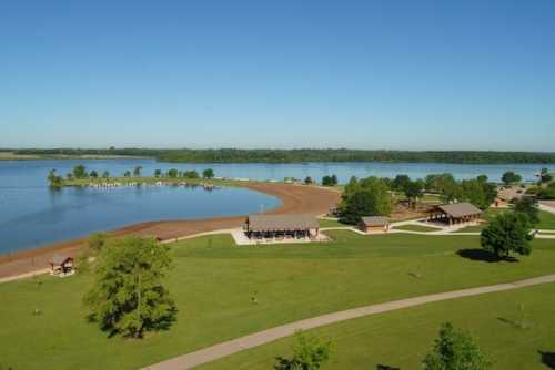 A scenic view of a lake with sandy shores, green lawns, and picnic pavilions under a clear blue sky.