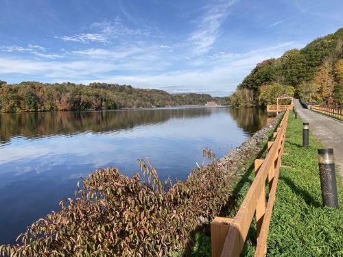 A serene lake surrounded by trees in autumn colors, with a walking path and wooden fence along the shore.