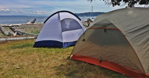 Two camping tents set up on grassy shore, with a view of the ocean and distant mountains under a clear blue sky.