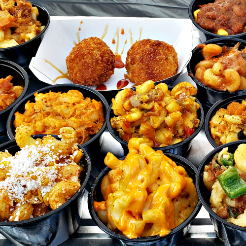 A variety of colorful dishes in black containers, including mac and cheese, croquettes, and pasta with vegetables.