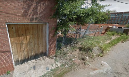 A wooden door on a brick wall next to a construction site, with a chain-link fence and overgrown grass nearby.