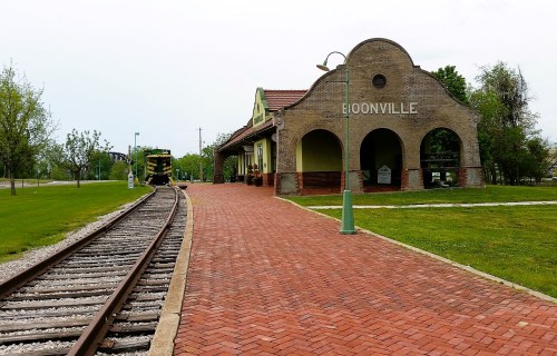 Historic train station in Boonville with a brick pathway and railway tracks, surrounded by green grass and trees.