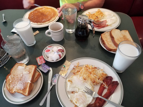 A breakfast spread featuring pancakes, eggs, bacon, toast, hash browns, milk, and coffee on a green table.