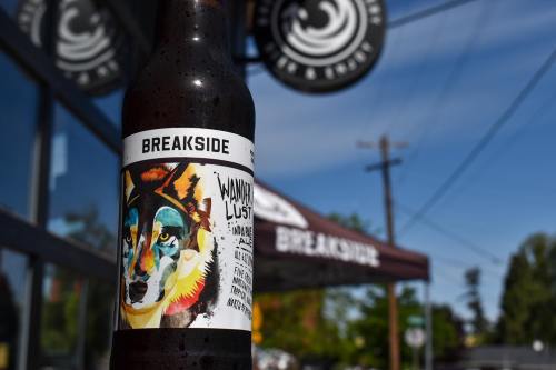 A close-up of a Breakside beer bottle featuring colorful wolf artwork, with a brewery sign in the background.