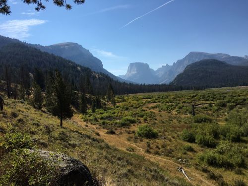 A scenic view of a valley surrounded by mountains under a clear blue sky, with green grass and trees in the foreground.