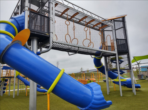 Colorful playground structure with blue slides, climbing features, and monkey bars under a cloudy sky.