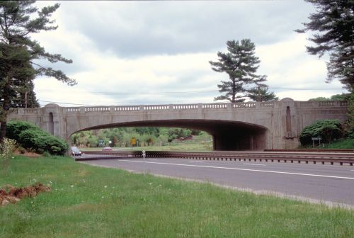A concrete bridge spans over a highway, surrounded by greenery and trees under a cloudy sky.