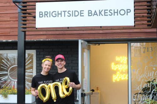 A couple stands outside Brightside Bakeshop, holding a gold "one" balloon, celebrating their first anniversary.