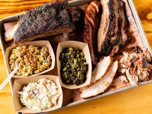 A tray of barbecue featuring ribs, brisket, sausage, coleslaw, greens, and popcorn, all served in paper containers.