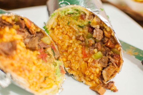 Close-up of a burrito cut in half, revealing a filling of rice, vegetables, and meat, wrapped in foil.