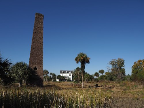 A tall, brick chimney stands beside a white house, surrounded by palm trees and open land under a clear blue sky.
