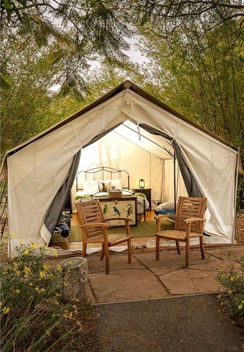 A cozy tent with a bed and chairs outside, surrounded by greenery and soft lighting.