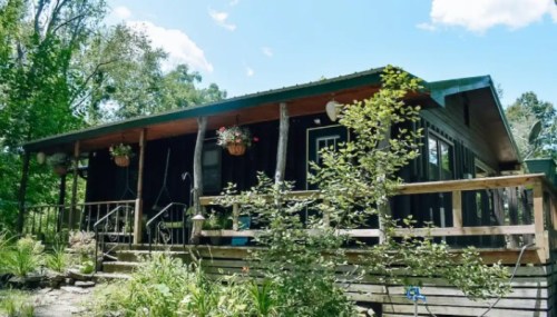 A cozy black cabin surrounded by greenery, featuring a wooden deck and hanging flower pots under a clear blue sky.